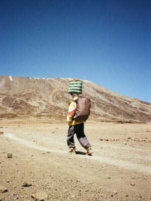 A young porter carries goods on a trek up Mount Kilimanjaro under a clear blue sky.