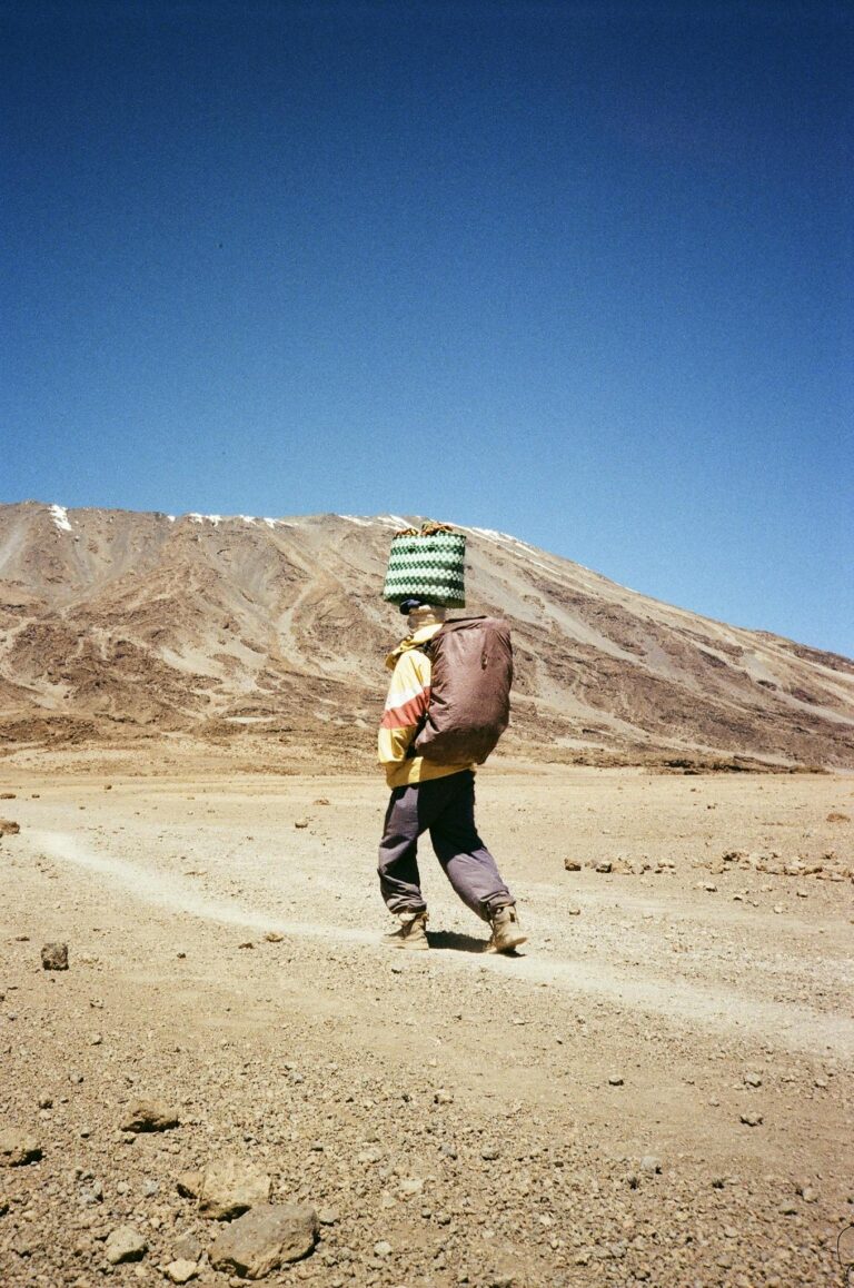 A young porter carries goods on a trek up Mount Kilimanjaro under a clear blue sky.
